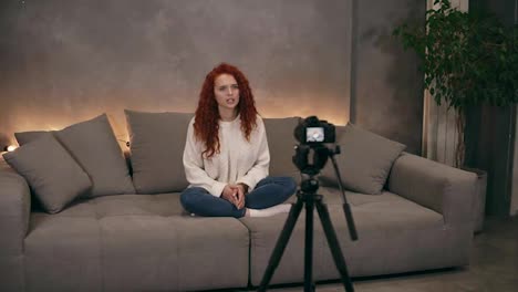 Portrait-of-long-haired-beautiful-woman-sitting-on-a-grey-big-couch-with-backlights-on-the-background-in-loft-interior-living-room-and-speaking-to-the-camera.-Young-blogger-making-a-video