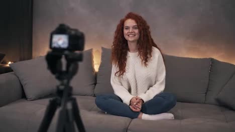 Attractive-curly-red-headed-young-girl-vlogger-is-talking-in-front-of-camera-recording-video-for-online-blog-in-internet-speaking,-smiling-and-gesturing.-Woman-is-wearing-jeans-and-white-sweater.-Accelerated-video