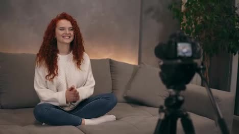 Caucasian-red-headed-young-girl-vlogger-is-talking-in-front-of-camera-recording-video-for-online-blog-for-her-followers,-smiling-and-gesturing.-Woman-is-wearing-jeans-and-white-sweater.-Accelerated-video.-Camera-on-foreground