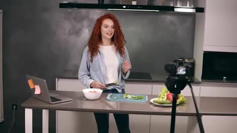 Young-woman-in-casual-clothes-leads-a-culinary-blog-and-records-video-in-grey-kitchen's-bar.-Video-shooting-of-the-blog---red-headed-cutting-tomatos-on-a-dashboard-and-explaining-a-recipe.-Modern-marketing,-social-networks