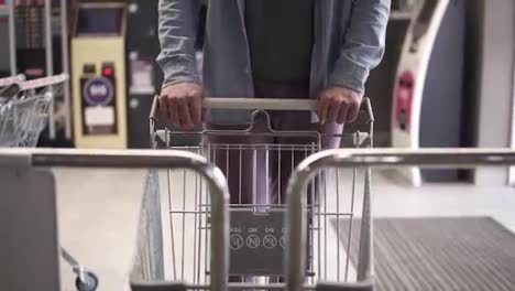 Cropped-front-footage-of-a-man-in-blue-shirt-and-violet-pants-pushing-the-shopping-empty-shopping-trolley-the-supermarket-entry.-The-guy-came-for-shopping-to-a-big-mall