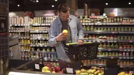 Portrait-of-young,-positive-man-shopping-for-fruits-and-vegetables-in-produce-department-of-a-grocery-store-supermarket.-Taking-oranges-and-putting-them-into-black-hand-carry-basket