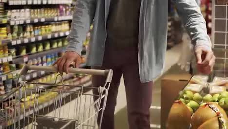 Cropped-footage-of-unrecognizable-man-in-blue-shirt-shopping-for-fruits-and-vegetables-in-produce-department-of-a-grocery-store-supermarket.-Taking-pomelo-fruit,-putting-it-into-shopping-trolley-and-walk-further.-Big-grocery-shelves-on-the-background