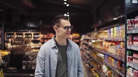A-handsome-young-caucasian-guy-in-glasses-and-blue-shirt-doing-shopping-in-the-supermarket,-walking-by-aisle,-looks-to-the-sides-and-smiling-cheerfully.-Slow-motion