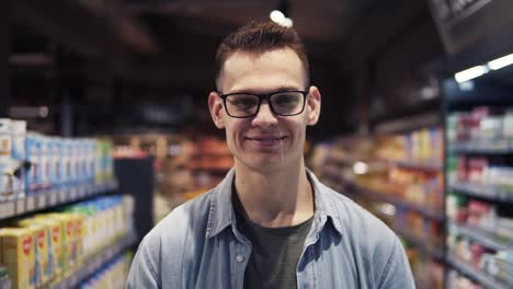 Close-up-of-the-handsome-young-caucasian-guy-in-glasses-doing-shopping-in-the-supermarket-and-smiling-cheerfully-to-the-camera.-Portrait
