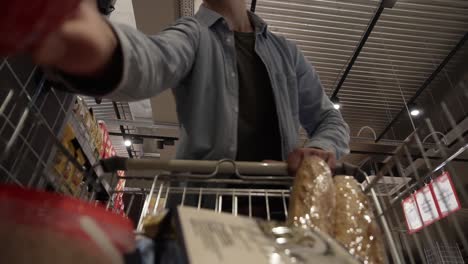 A-caucasian-young-guy-in-glasses-putting-goods-in-shopping-trolley.-Passing-through-the-grocery-shelves-of-a-big-supermarket-and-reading-labels.-Low-angle-footage-from-the-moving-shopping-cart
