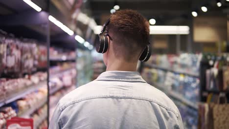 At-the-Supermarket:-Stylish-caucasian-guy-with-headphones-walks-through-meat-goods-section-of-the-store.-Following-Back-view-shot.-Slow-Motion.-Blurred-background