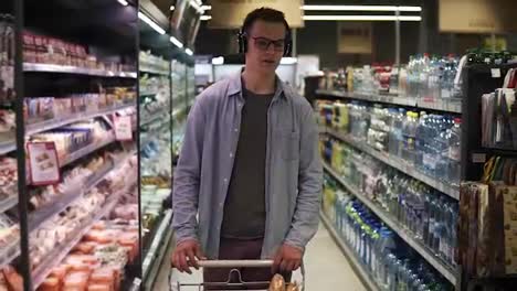 Attractive-short-haired-guy-in-stylish-glasses-listening-to-the-music-while-doing-shopping-in-a-big-gtocery-store.-Passing-by-aisels-with-goods,-pushing-cart.-Front-view
