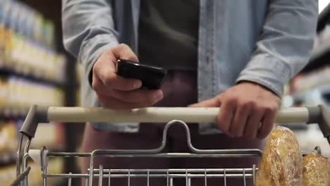 Cropped-close-up-footage-of-a-young-man-pushing-trolley-cart-against-blurred-background-and-shopping-in-grocery-supermarket.-Concentrated-male-using-modern-smartphone-and-choosing-fresh-food