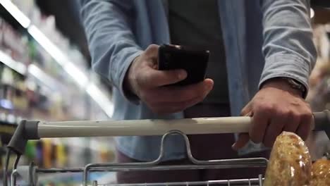 Cropped-close-up-footage-of-a-young-man's-hands-with-stylish-watches-pushing-trolley-cart-against-blurred-background-and-shopping-in-grocery-supermarket.-A-guy-using-modern-smartphone-and-choosing-fresh-food
