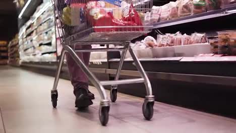 Shopping-in-the-supermarket,-view-of-the-male-legs-from-inside-the-trolley.-A-guy-pushing-full-of-goods-shopping-trolley-by-the-supermarket-aisle.-Cropped,-close-up-footage