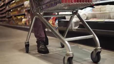 Shopping-in-the-supermarket,-view-of-the-male-legs-from-inside-the-trolley.-A-guy-pushing-shopping-cart-by-the-supermarket-aisle.-Cropped,-close-up-footage.-Slow-motion