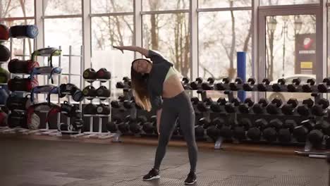 Full-length-shot-of-determined-fitness-woman-warming-up-in-modern-gym.-Muscular-sportswoman-warming-up-before-a-intense-workout-standing-in-front-the-wall-of-dumbbells-and-bright-window