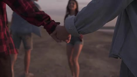 Close-up-footage-of-a-couple-holding-hands-together-while-walking-on-the-beach.-Another-couple-on-a-perspective.-Young-people-having-fun-together.-Dusk