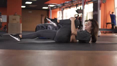 Side-view-of-a-lying-on-back-woman-performs-exercise-of-twisting-the-body-for-flexibility.-Close-up-of-a-female-person-in-a-grey-elastic-sportswear-makes-gymnastics-on-floor,-stretching-her-legs