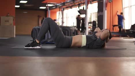 Young-european-woman-in-grey-sportswear-pumps-the-abdominal-muscles-on-the-floor-in-the-modern-gym.-Sport-and-fitness-concept.-Footage-from-the-side.-Slow-motion