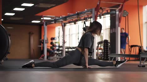 Flexible-skinny-girl-stretches-before-the-split,-doing-the-split,-stretching-to-left-leg-and-makes-a-slope-forward-on-the-mat-in-the-modern,-stylish-gym.-Side-view