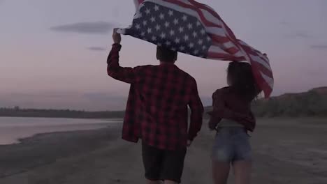 Young-couple-both-in-red-plaid-shirts-are-running-on-the-seashore-with-american-flag-raised-above-their-heads.-Backside-view