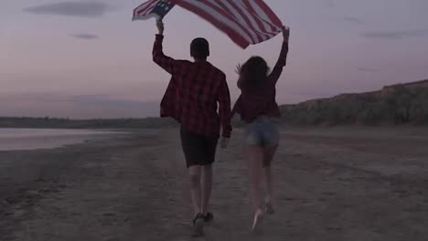 Young-woman-and-man-running-on-the-sand-with-raised-american-flag,-feeling-freedom.-Evening-dusk.-Tracking-footage