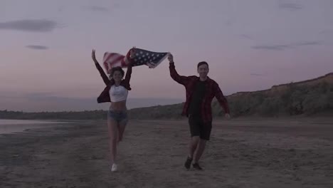Beautiful-young-two-people-running-on-a-seaside-in-the-evening.-dusk-holding-above-an-american-flag.-Happy,-smiling,-emotions