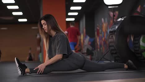Flexible-long-haired-european-woman-working-on-extreme-leg-stretch-splits-exercise-im-modern,-crossfit-gym.-Determinated-girl-in-sportswear-working-on-her-flexibility.-Side-view