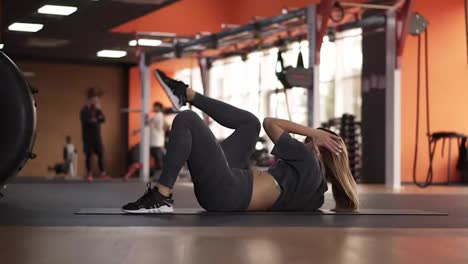 Concenrated-young-woman-in-grey-sportswear-pumps-the-abdominal-muscles-on-the-floor-in-the-modern-gym,-bending-up-to-the-legs-with-hands.-Sport-and-fitness-concept.-Footage-from-the-side.-Slow-motion