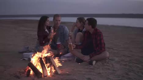 Carefree-young-friends-spending-time-together-And-drinking-beer-by-bonefire-on-the-beach-as-the-sun-begins-to-set.-Talking,-discussing-something