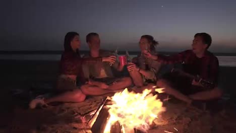 Group-of-people-are-spending-time-near-the-bonfire-on-the-beach-at-night.-Drinking-alcohol,-cheers.-Young-man-is-holding-a-guitar.-Front-view