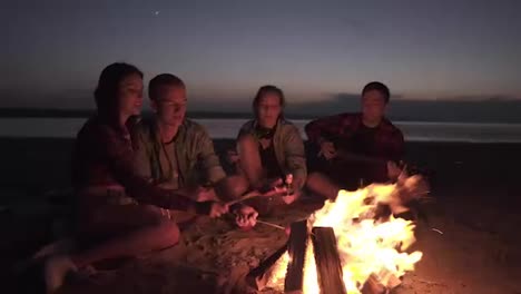 Picnic-with-friend-on-the-beach-near-the-bonfire.Young-people-are-roasting-sausages-on-wooden-sticks.-Male-from-the-side-is-playing-the-guitar.-Night-time
