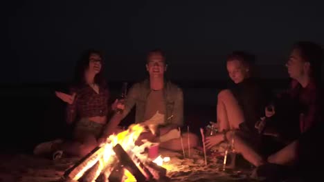 Group-of-friends-hanging-out-together-on-the-beach.-Beautiful-two-couples.-Singing-song-with-guitar,-gesturing.-Friendship-concept.-Bonfire-at-night