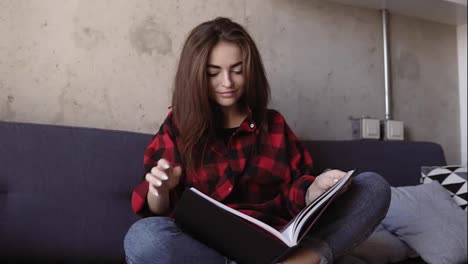 Attractive-young-brunette-girl-in-comfy-red-flannel-shirt-sitting-on-a-couch-and-reading-a-book.-Chilling-out-time.-Slomotion-footage.