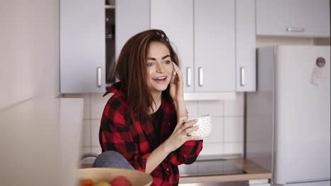 Young-beautiful-girl-starts-calling-someone-while-sitting-in-her-kitchen-and-having-a-cup-of-tea-in-her-hand.
