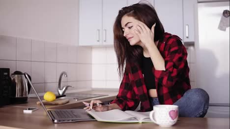 Attractive-girl-in-her-20's-looks-forsomething-on-the-internet-using-her-laptop-and-then-writes-something-down-in-her-notebook-while-sitting-in-a-cozy-kitchen-of-her-apartment