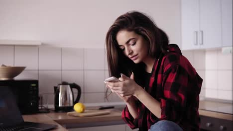 Young-beautiful-brunette-girl-with-messy-hair-in-flannel-shirt-sitting-in-her-kitchen-and-texting-someone.