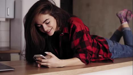 Attarctive-brunette-girl-is-dreaming-and-smiling-about-something-while-lying-on-kitchen-table-surface-and-texting-someone.
