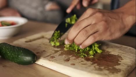 Close-up-slow-motion-footage-of-a-young-mulatto-guy-cutting-lettuce-for-salad,-making-lunch-with-his-girlfriend-who's-sitting-beside-him-on-the-kitchen-table-surface.