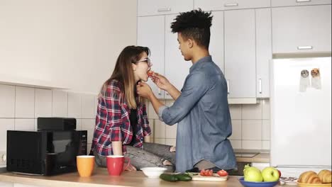 Two-young-people-enjoying-their-time-together,-making-lunch-in-comfy-kitchen.-Living-together.-Mulatto-guy-is-feeding-slice-of-tomato-to-his-attractive-caucasian-girlfriend.
