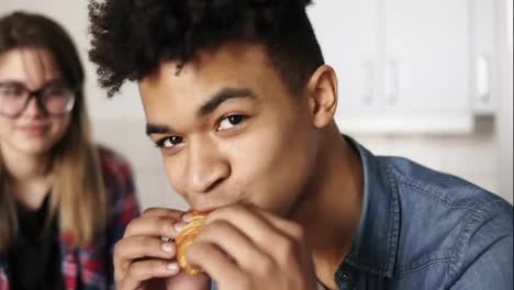 Close-up-footage-of-a-handsome-mulatto-guy-in-his-20's-with-expressive-deep-dark-brown-eyes-looking-at-the-camera,-eating-croissant,-with-his-girlfriend-sitting-in-the-background.