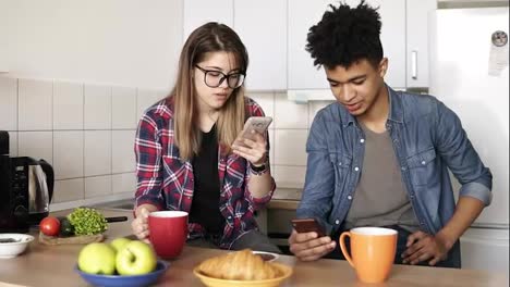 Cute-couple-having-breakfast-together-at-home-in-their-kitchen,-attractive-caucasian-girl-and-her-mulatto-boyfriend-using-their-smartphones,-not-talking-to-each-other-at-first.