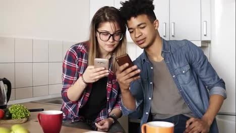 Attractive-guy-and-a-girl-both-in-their-20's-scrolling-their-smartphones,-exchanging-information-while-sitting-in-a-comfy-spacious-kitchen.