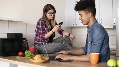 Junges,-Glückliches-Hipsterpaar,-Das-In-Der-Küche-Sitzt-Und-Beide-Geräte-Wie-Laptop-Oder-Smartphone-Benutzt,-Ohne-Miteinander-Zu-Reden.