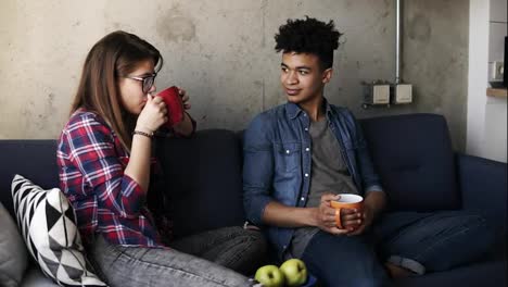 Cute-young-couple,-attractive-caucasian-girl-and-her-handsome-mulatto-boyfriend-sitting-on-the-couch,-drinking-tea,-enjoying-their-time-together.