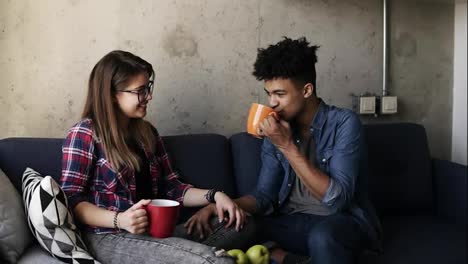 Young-caucasian-girl-and-handsome-mulatto-guy-both-wearing-urban-outfits,-sitting-on-the-couch-with-cups-of-tea,-talking-about-life,-enjoying-spending-time-together.