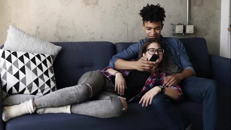 Cute-young-couple-on-the-couch,-attractive-caucasian-girl-lying-on-her-handsome-mulatto-boyfriend's-laps.-He-is-trying-to-find-a-perfect-tv-chanel,-with-the-remote-in-his-hand.