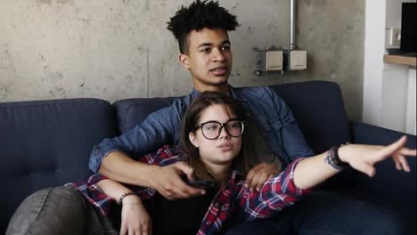 Cute-happy-couple-of-young-hipsters-lying-on-the-couch,-trying-to-find-a-perfect-tv-channel.-Leisure-time,-enjoying-youth,-living-together.-Relationship-goals.