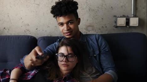 Cute-happy-couple-of-young-hipsters-lying-on-the-couch,-laughing-while-watchingsomething-on-TV.-Leisure-time,-enjoying-youth,-living-together.-Relationship-goals.