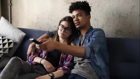 Cute-happy-couple-of-young-hipsters-cuddling-on-the-couch,-trying-to-find-a-perfect-tv-channel.-Leisure-time,-enjoying-youth,-living-together.-Relationship-goals.