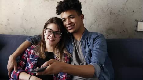 Cute-happy-couple-of-young-hipsters-smiling-and-cuddling-on-the-couch-while-trying-to-find-a-perfect-tv-channel.-Leisure-time,-enjoying-youth,-living-together.-Relationship-goals.