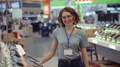 Positive-female-seller-or-shop-assistant-portrait-in-supermarket-store.-Woman-in-blue-shirt-and-empty-badge-looking-at-the-camera-and-smiling.-Electronic-devices-on-the-background.-Slow-motion