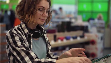 Curly-woman-with-headphones-on-neck-standing-at-the-counter-with-mobile-phones-in-casual-clothes-choosing-a-new-smartphone-in-a-modern-electronics-store